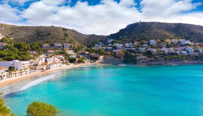 Bahía de aguas turquesas en cala el Portet, con casas blancas y verdes colinas.