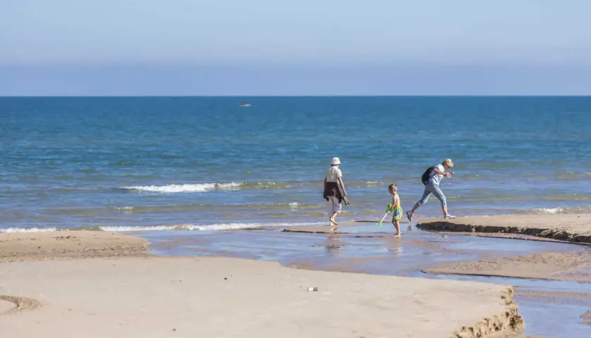 Un niño y dos adultos disfrutan de un día soleado en la playa Pau Pi, jugando cerca de la orilla del mar azul.