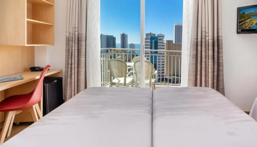 Habitación de hotel pueblo Benidorm con vistas al mar y rascacielos. Balcón con mesa y sillas. Escritorio con silla roja y minibar.