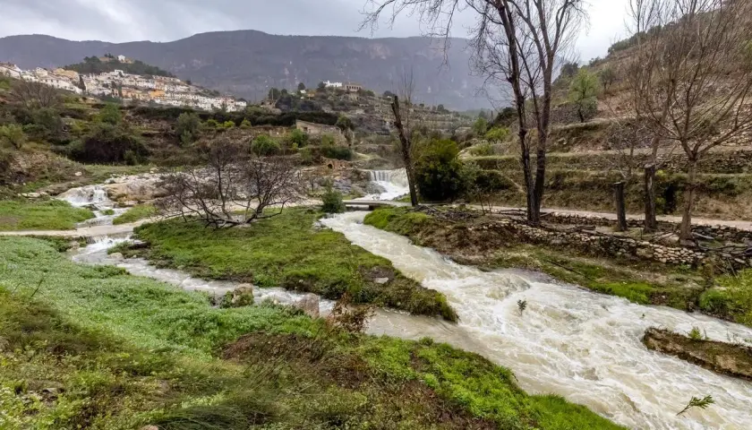 Río caudaloso cerca de Sella, Alicante, con cascadas y un pueblo blanco en la ladera.