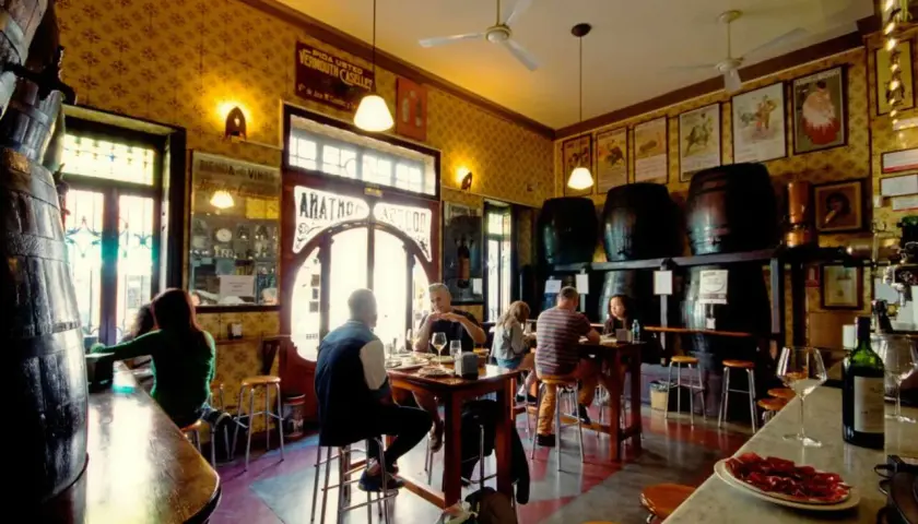 Gente en un bar tradicional, con barriles y carteles antiguos. Un lugar con encanto, como una casa de montaña en Valencia, donde disfrutar de tapas y vermut.