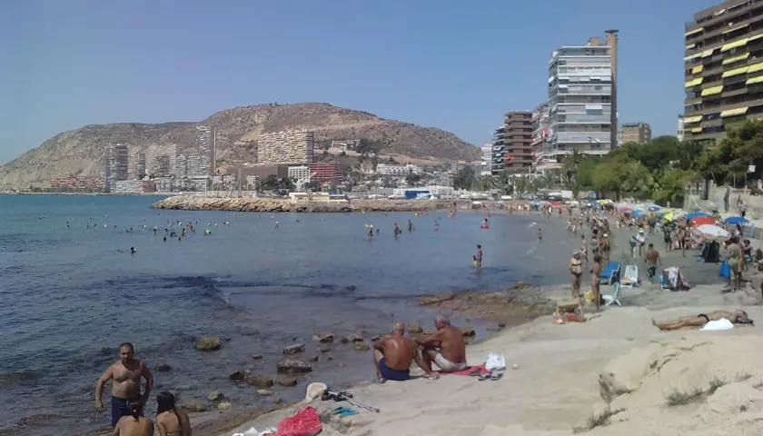 Gente disfrutando del sol y el mar en la playa de la Almadraba, con edificios modernos al fondo.