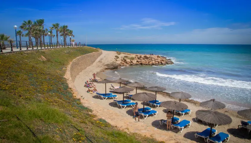 Una cala estaca con palmeras y sombrillas en la arena, donde la gente descansa bajo el sol. El mar azul turquesa invita a un refrescante baño.