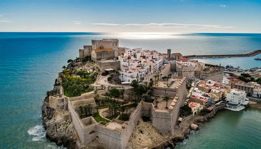 Castillo de Peñíscola sobre el mar, con sus murallas y casas blancas. La peñíscola playa se extiende a su lado, invitando a disfrutar del sol y el Mediterráneo.