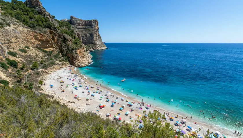 La **cala Moraig** es un paraíso de guijarros y aguas turquesas, con acantilados imponentes y gente disfrutando del sol bajo sombrillas de colores.