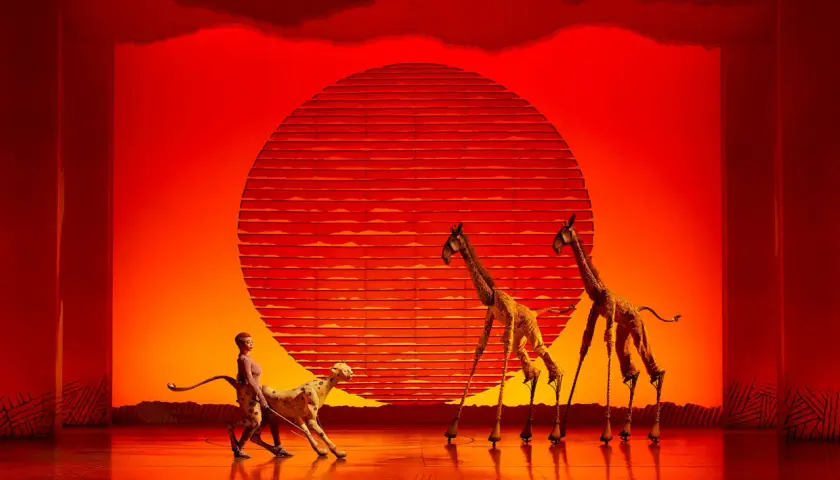 Escena de teatro en Valencia con un guepardo y jirafas con zancos ante un sol naranja.
