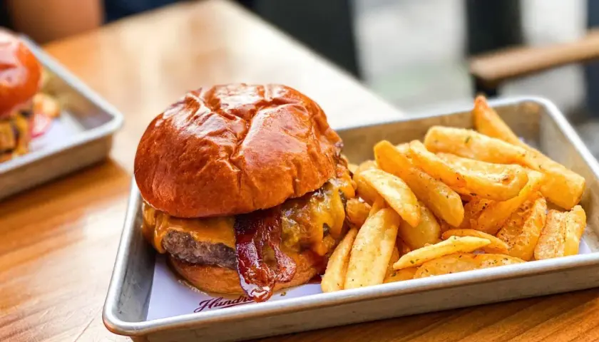 Jugosa hamburguesa con queso fundido y tocino crujiente, acompañada de patatas fritas doradas. Un festín para los sentidos en Hundred Valencia.