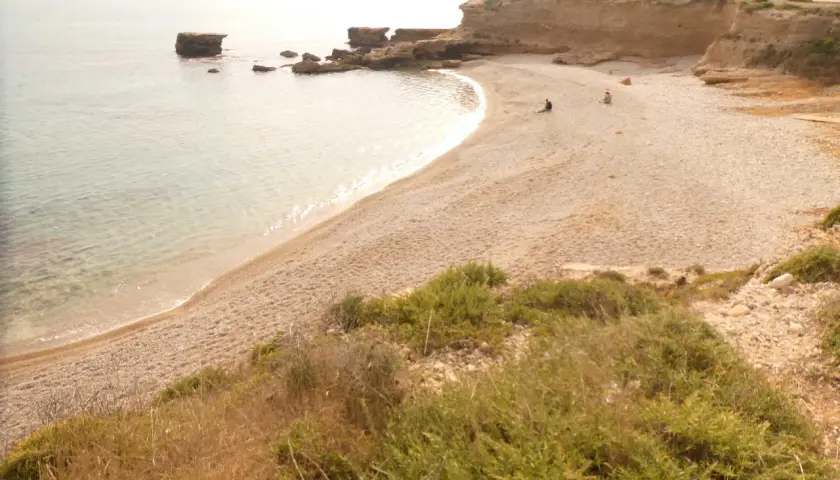 Una tranquila cala foradada con aguas cristalinas y rocas emergiendo del mar. Dos personas disfrutan de la soledad en la playa de guijarros.