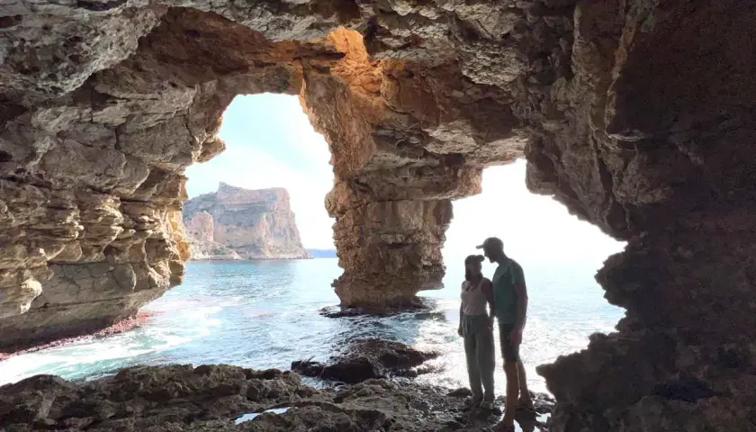 Pareja contemplando el mar desde la Cova dels Arcs, con sus impresionantes formaciones rocosas y el sol filtrándose.