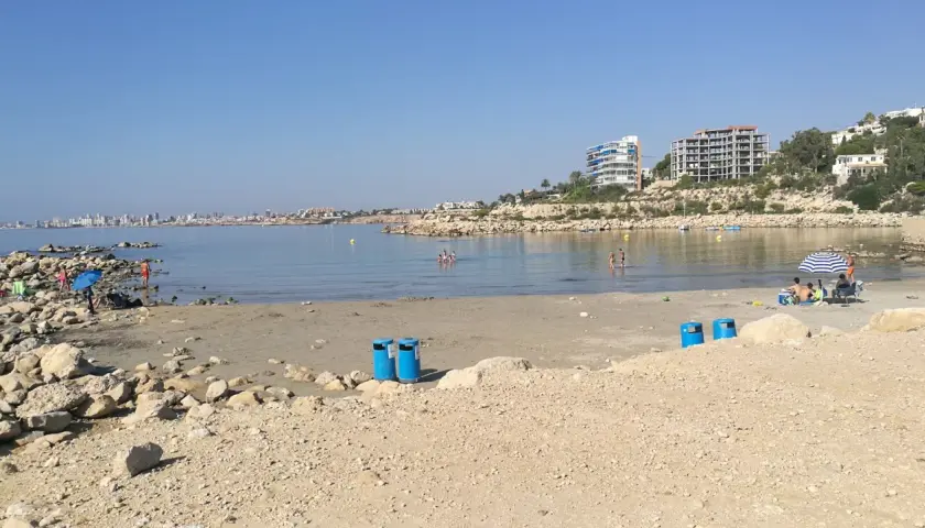 Día soleado en la cala Morro Blanc. Gente disfrutando del mar y la playa, con edificios modernos al fondo.