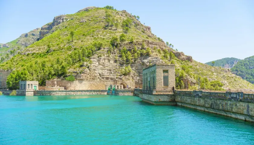 El embalse de Benagéber, con sus aguas turquesas y la imponente presa de piedra, se funde con la montaña verde.