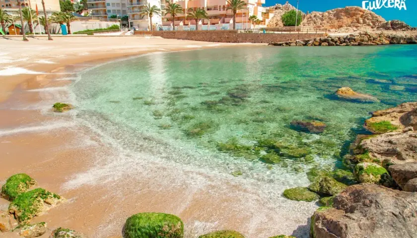 Aguas cristalinas y arena dorada en la cullera playa, con edificios y palmeras al fondo.