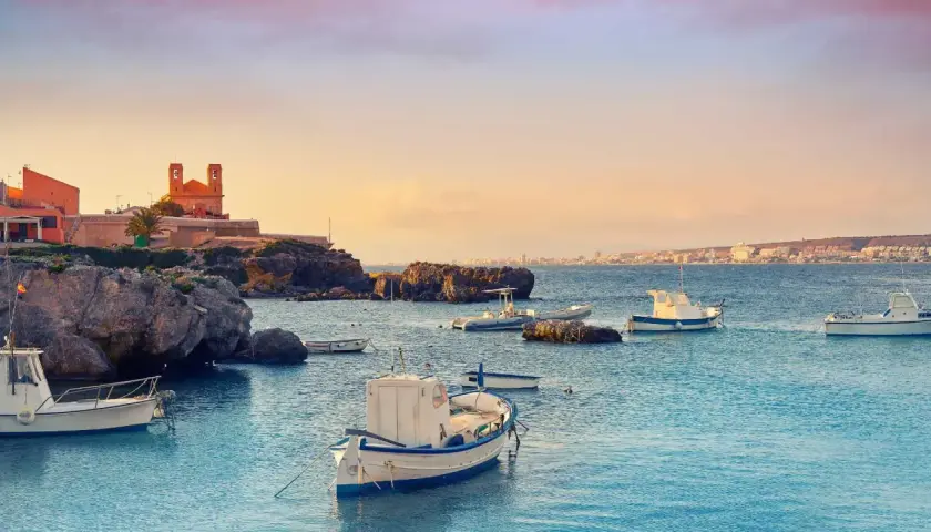 Barcas tranquilas en el puerto Santa Pola, con edificios históricos y la costa al fondo bajo un cielo anaranjado.
