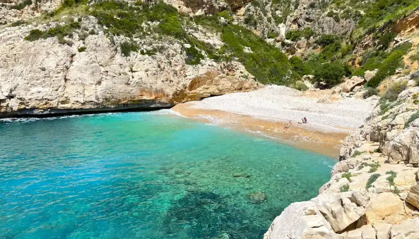 Aguas turquesas bañan la cala testos, una playa de guijarros rodeada de acantilados rocosos y vegetación.