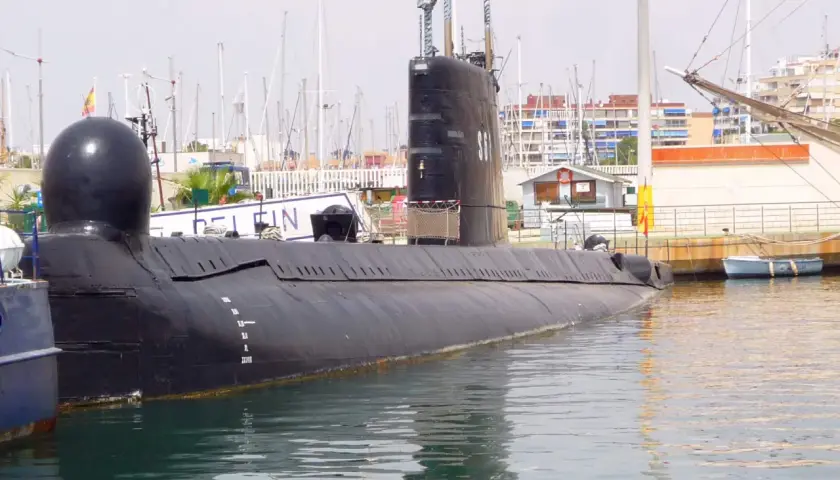 Un submarino negro, el Delfín, descansa en el puerto de Torrevieja, con mástiles de veleros y edificios al fondo.