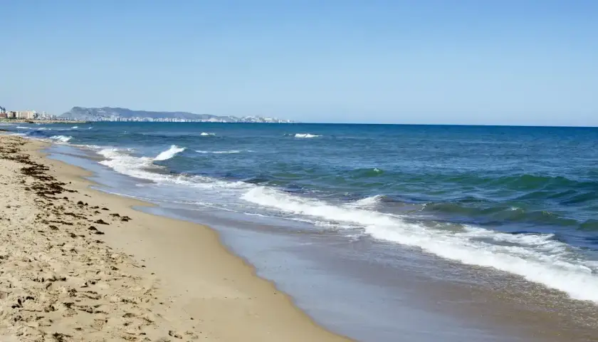 La playa de Xeraco, con arena dorada y el mar azul, se extiende hacia un horizonte con edificios y montañas.