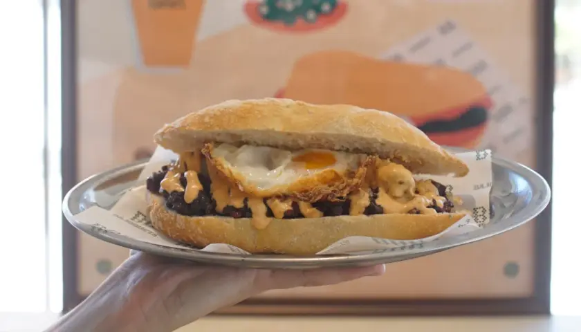 Un delicioso almuerzo valenciano: un bocadillo con carne, huevo frito y salsa cremosa, servido en plato metálico.