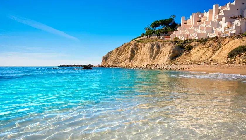 Aguas cristalinas en las calas de Alicante, con una urbanización blanca en la cima del acantilado y el mar azul intenso.