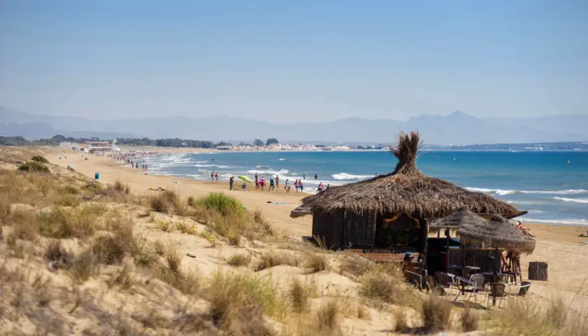 Chiringuito de paja en la playa del Rebollo, con gente disfrutando del sol y el mar azul. Montañas al fondo.