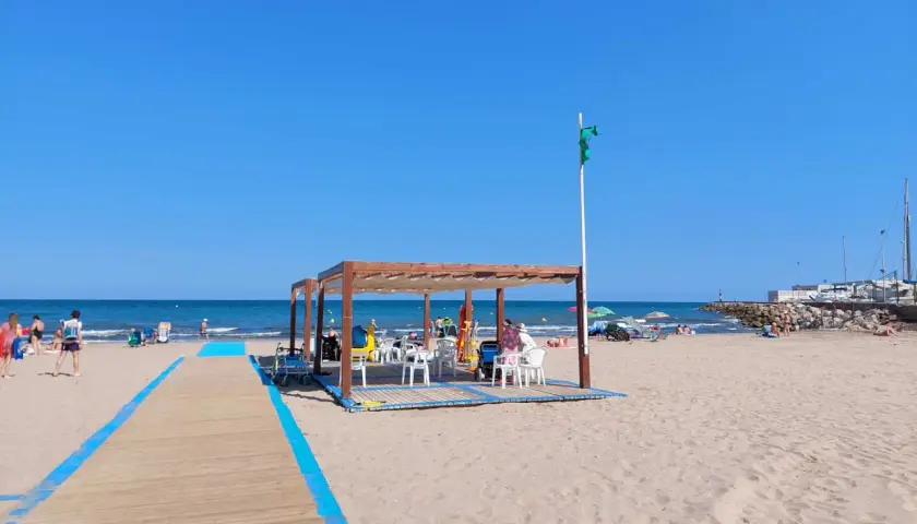 Pasarela de madera azul en la playa de Massamagrell, con chiringuito y gente disfrutando del sol y el mar.