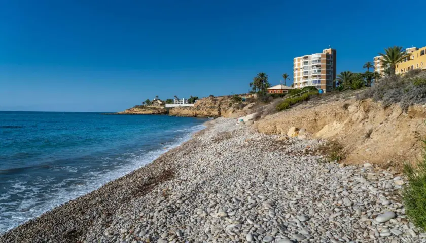 La cala Amerador, con su playa de guijarros y aguas turquesas, se extiende bajo un cielo azul. Edificios modernos y vegetación mediterránea bordean la costa.