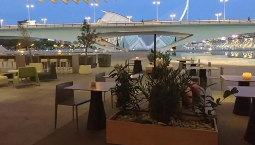 Mesas y sillas en la terraza de restaurantes con vistas a un puente moderno y un cielo azul crepuscular.