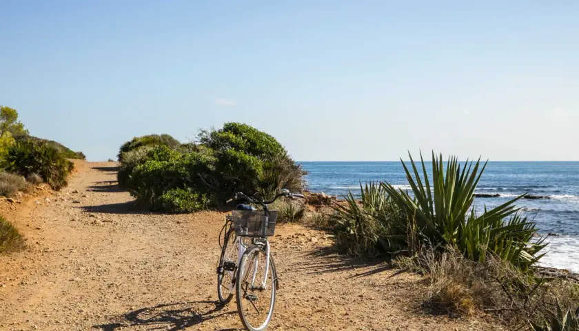 Bicicleta blanca en un camino de tierra junto al mar. El sol ilumina la escena, creando sombras alargadas. Parece que la renega ha dejado su bici para disfrutar de la brisa marina.