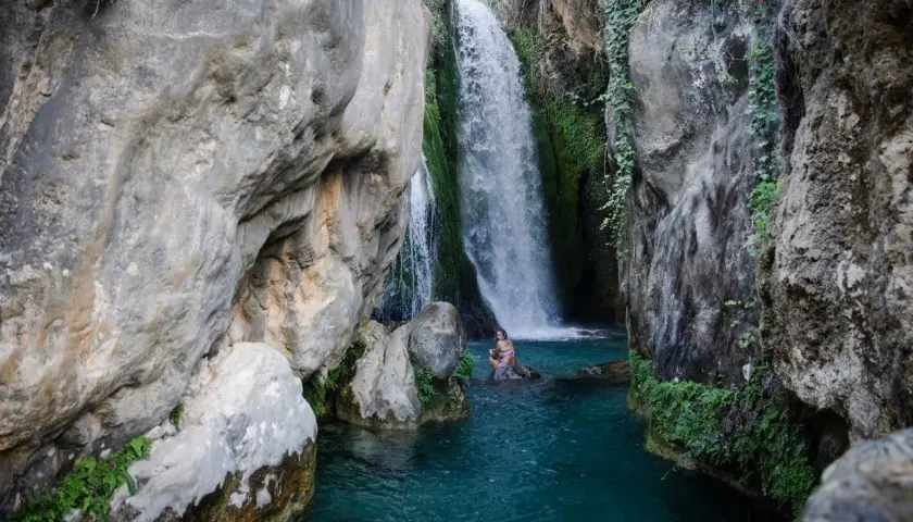 Una mujer se baña en las **fonts de Algar**, rodeada de rocas y vegetación, bajo una cascada de agua cristalina.