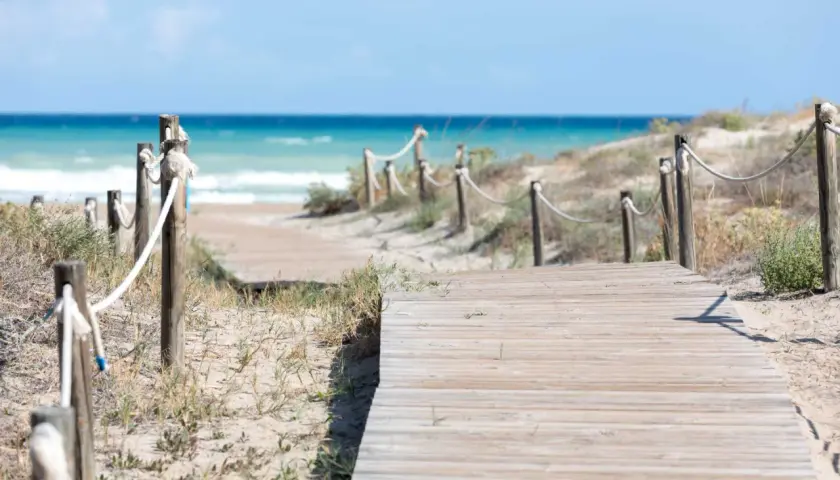 Pasarela de madera que conduce a la playa Raco de la Mar. El mar azul y las dunas de arena crean un paisaje sereno.