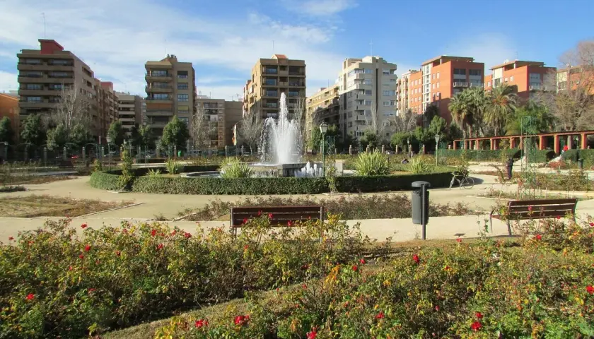 Parque urbano con fuente central y edificios residenciales al fondo. Los viveros de Valencia lucen sus rosales en flor.