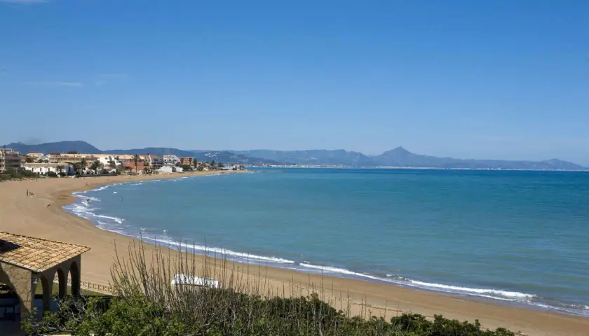 Amplia vista de la playa de Les Bovetes con arena dorada, mar azul y montañas al fondo. Un día soleado perfecto para disfrutar.