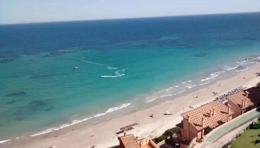 Vista aérea de la Campoamor playa con aguas turquesas, gente disfrutando del sol y un kitesurfista en acción.
