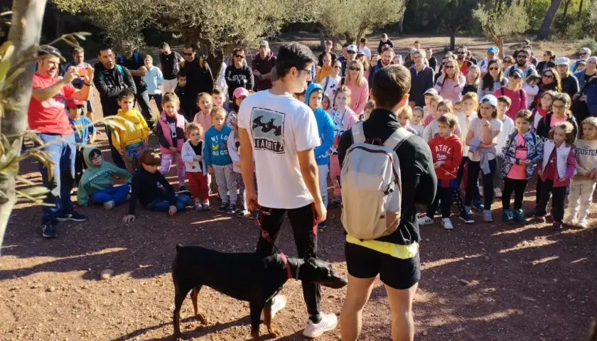 Un grupo de personas, incluyendo niños, se reúne en un sendero boscoso. Un hombre con una camiseta roja toma una foto con su móvil. Un perro Dóberman negro está atado a un joven. Quizás están buscando al **dragón de la Calderona**.