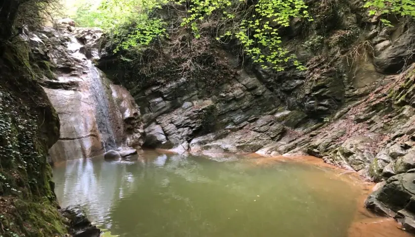 Un rincón secreto en el gorg dels Banyuts, con una cascada que alimenta una poza de aguas verdes rodeada de rocas y vegetación exuberante.