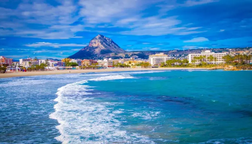 Olas turquesas rompen en la arena dorada de la playa del Arenal Javea, con edificios y el imponente Montgó al fondo.