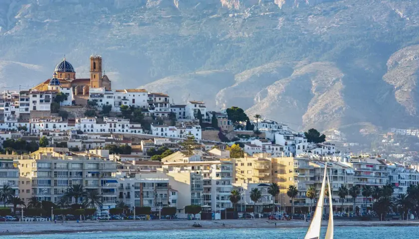 Vista del **castell de l'Olla** coronando la colina, con sus casas blancas y la iglesia de cúpula azul. Un velero navega frente a la costa.