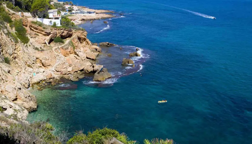 Vista de la cala Les Rotes con aguas cristalinas, acantilados rocosos y un kayak amarillo en el mar azul.