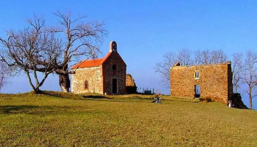 La ermita de Santa Catalina, una pequeña iglesia de piedra con tejado naranja, se alza junto a unas ruinas bajo un cielo azul. Un árbol desnudo y un paisaje verde completan la escena.