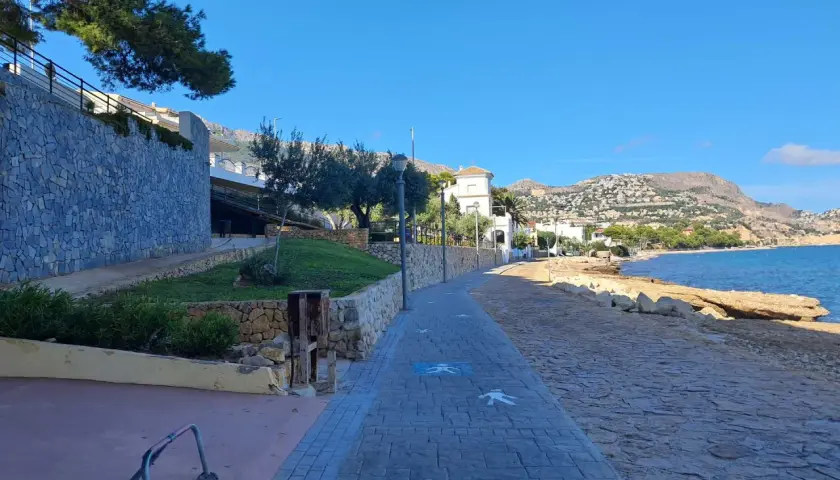 Paseo marítimo con siluetas de personas en la cala del Soio. El mar azul y las montañas de fondo crean un paisaje idílico.