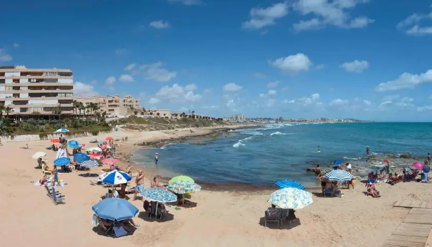 Gente disfrutando del sol y el mar en la playa Cabo Cervera, con sombrillas de colores y edificios al fondo.