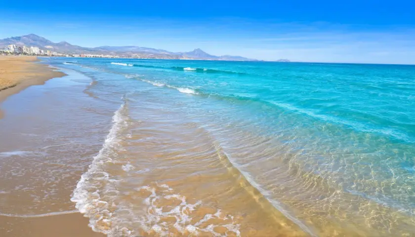 Aguas turquesas y arena dorada en las calas de San Juan, Alicante. El mar Mediterráneo acaricia la costa, con montañas al fondo.