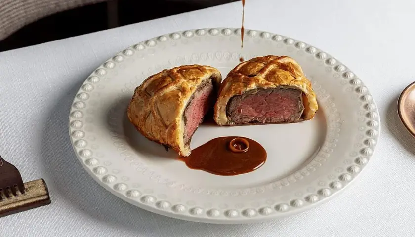 Un Wellington de ternera jugoso, cortado por la mitad, servido con salsa en un plato elegante. Una delicia para probar en los restaurantes de Valencia.