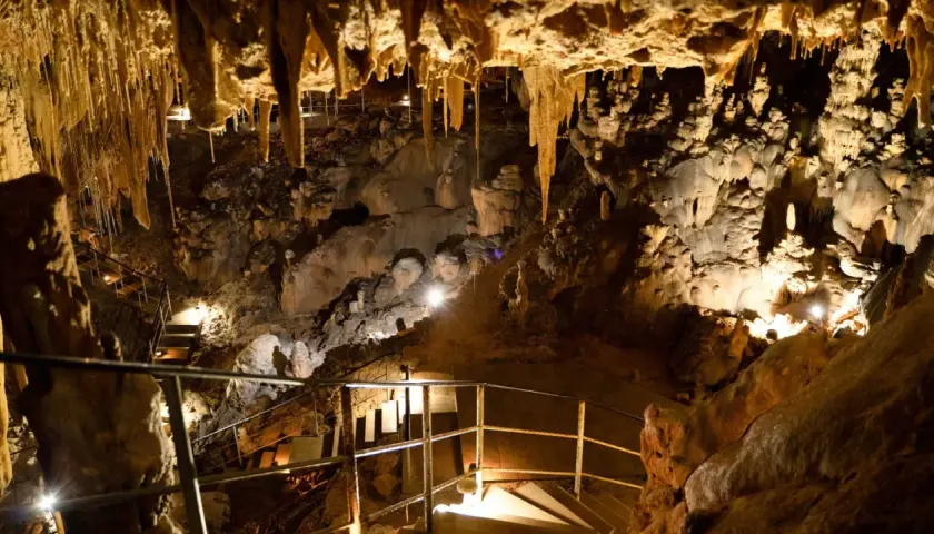 Interior de la cueva del Rull, con estalactitas colgando del techo y formaciones rocosas iluminadas. Escaleras y barandillas guían el recorrido.