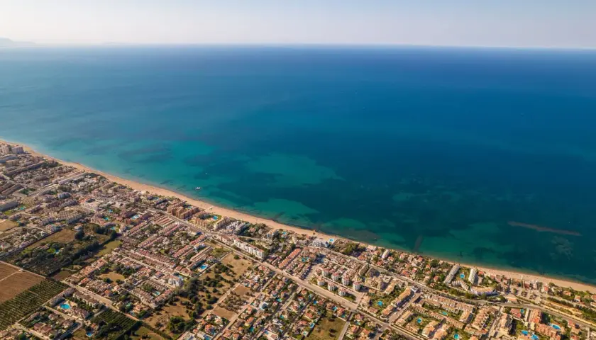 Vista aérea de la playa de arena de Dénia, con el mar azul turquesa y edificios residenciales.