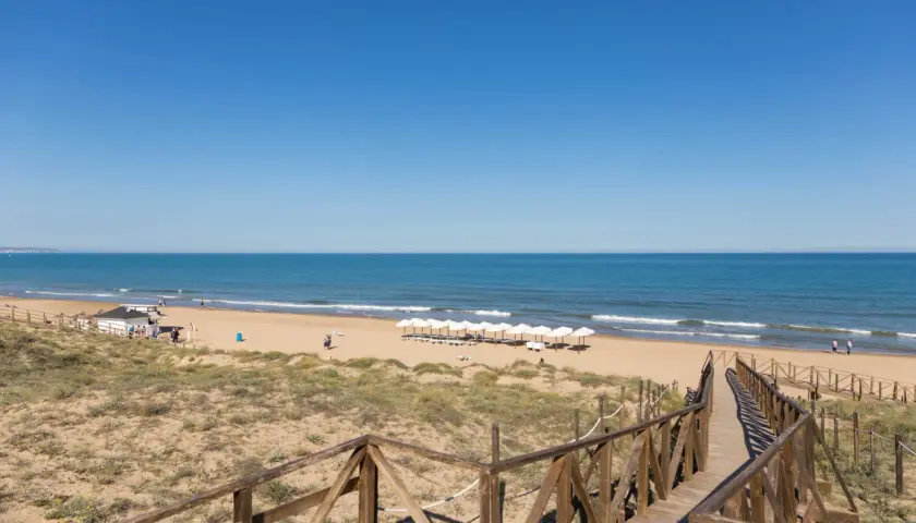 Pasarela de madera hacia la playa Moncayo, con arena dorada, mar azul y sombrillas blancas alineadas.