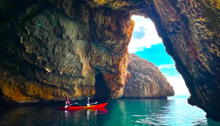 Dos personas en kayak rojo exploran la cueva de la cala del llop mari, con aguas turquesas y formaciones rocosas impresionantes.