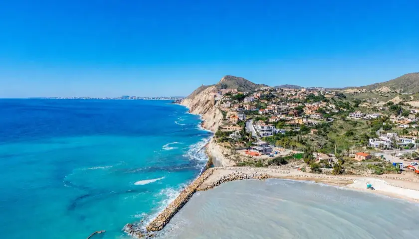 Vista aérea de la costa con casas blancas y el mar azul turquesa. La cala Baeza se extiende hacia el horizonte, con una bahía protegida por un rompeolas de rocas.