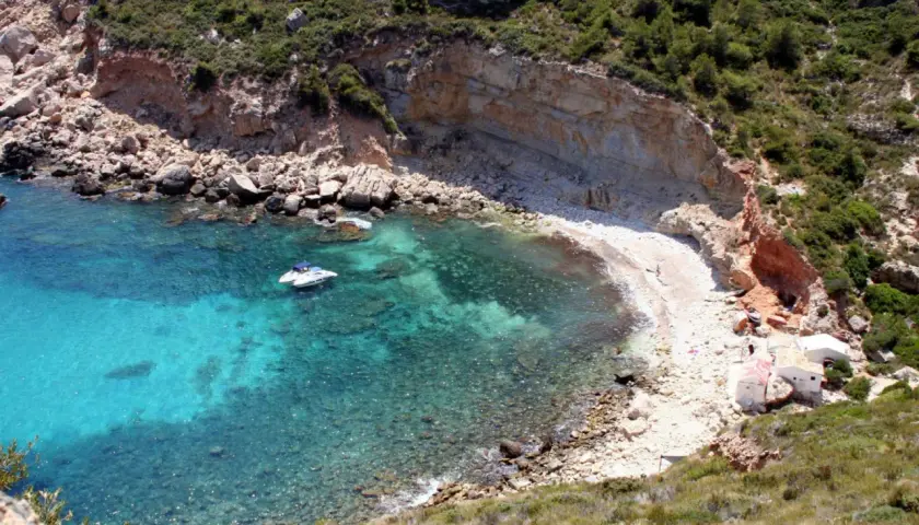 Aguas turquesas y una cala rocosa en Cala Llebeig, con un pequeño barco y casitas blancas en la orilla.