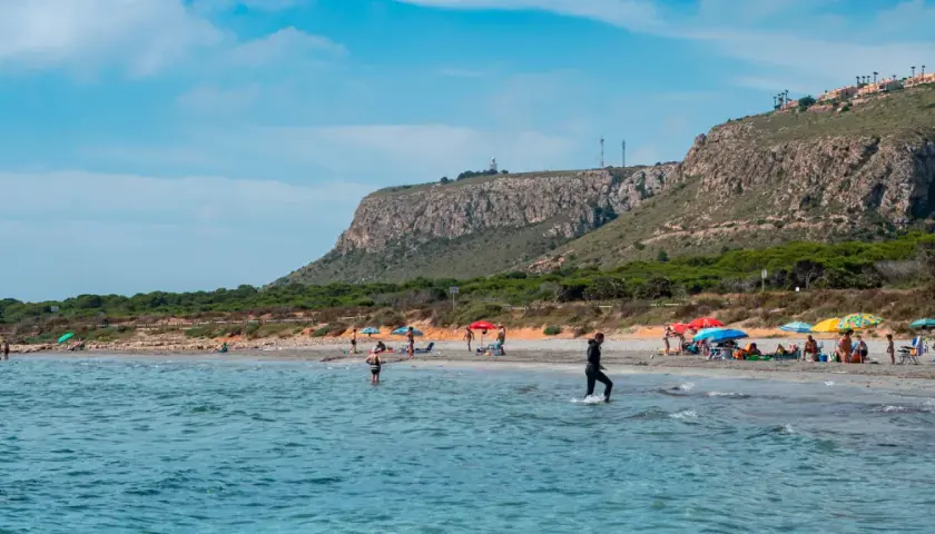 Gente disfruta del sol en la playa de la ermita santa pola, con aguas turquesas y un acantilado verde al fondo.