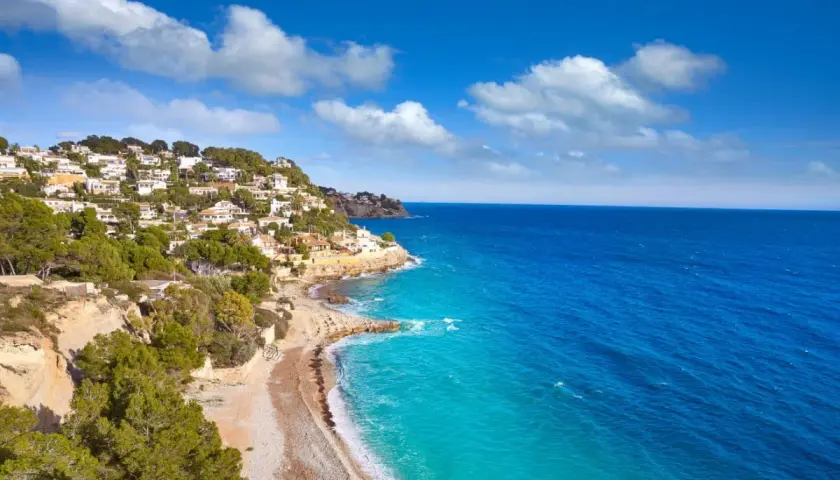 La **cala Baladrar** se asoma entre casas blancas y pinos verdes, con aguas turquesas y un mar azul intenso bajo un cielo nublado.